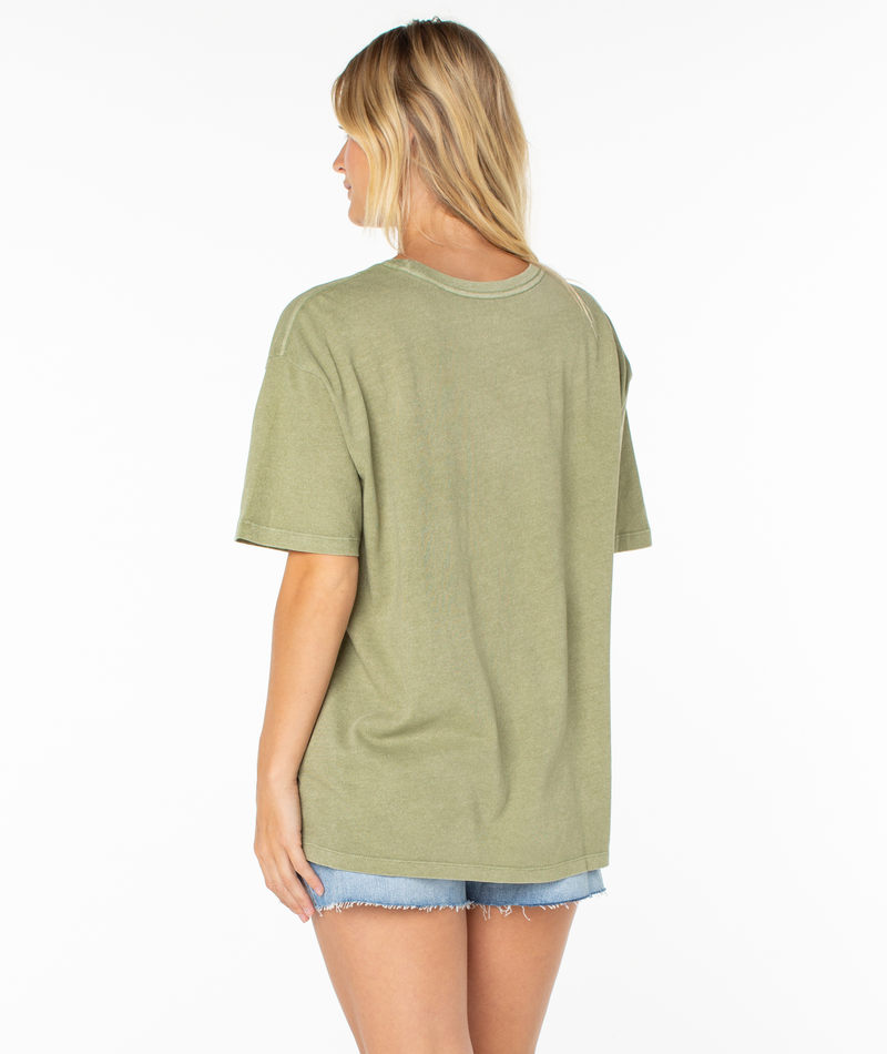 A person with long blonde hair stands facing away against a white background, wearing the Roxy Surfin Tee in olive green with a relaxed drop shoulder fit and light blue denim shorts.