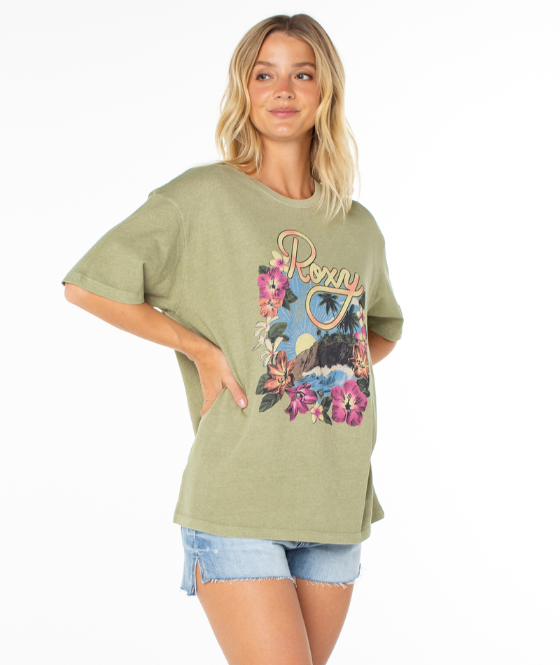 A woman wears the Roxy Surfin Tee, featuring a colorful surf and floral graphic, paired with light denim shorts. She smiles with one hand on her hip against a plain white background.
