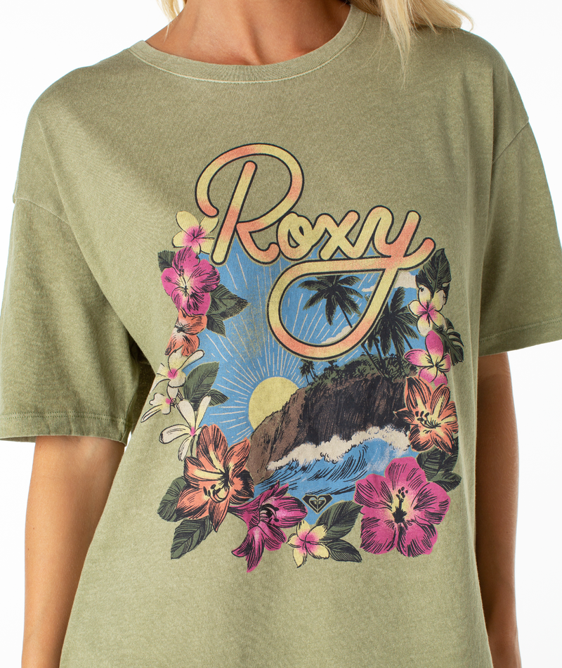 A person wears the Roxy Surfin Tee, an oversized top with a relaxed drop shoulder and a tropical graphic of flowers, sun, ocean waves, and rocky landscape, featuring the Roxy logo.