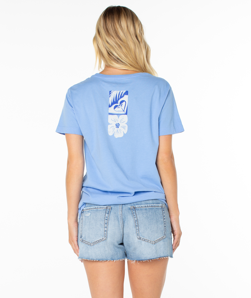 A woman with long blonde hair stands facing away from the camera in a Roxy Beach Things Tee, featuring a floral back graphic, and light denim shorts against a white background.