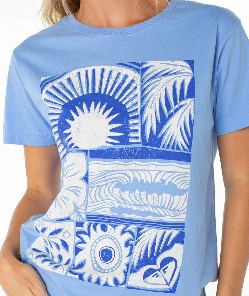 A person wearing the Roxy Beach Things Tee, a light blue top with bold white and blue abstract sun, wave, leaf, and heart graphics in rectangular panels.