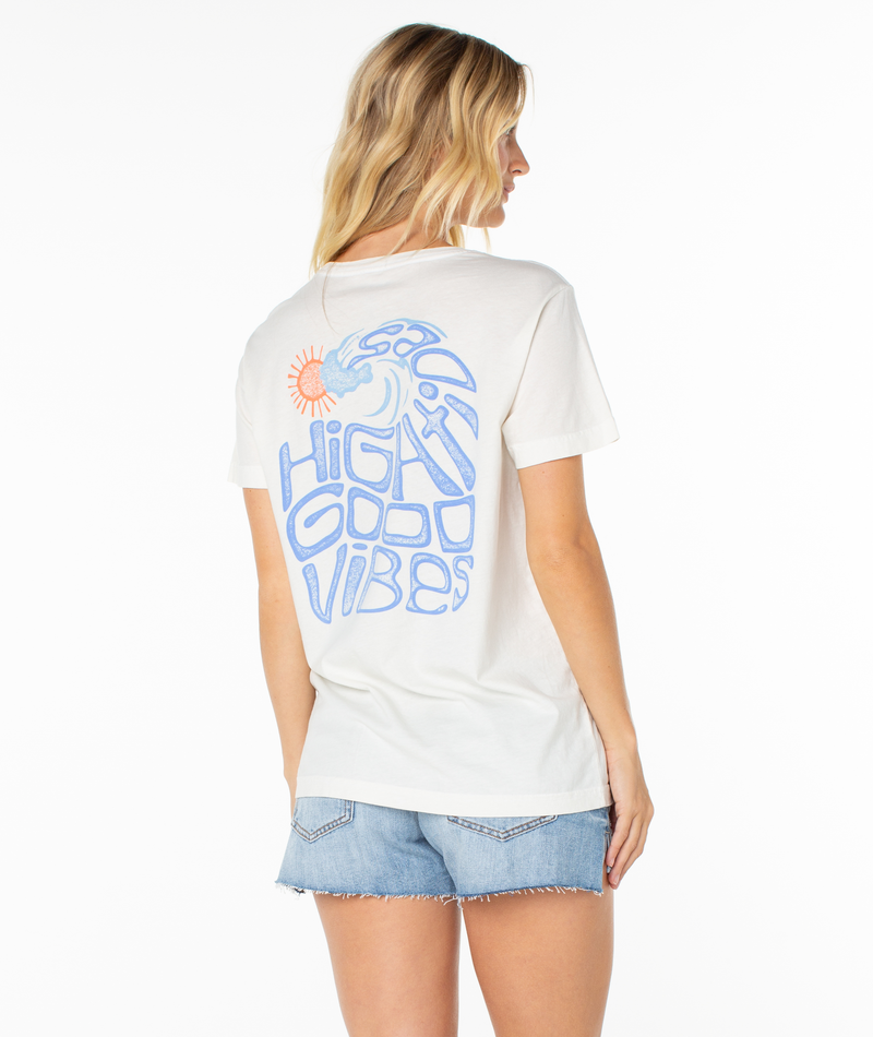 A woman with blonde hair is seen from behind wearing the Roxy High Tides Tee—a white beachy top with blue "HIGH GOOD VIBES" text and a sun and wave graphic on the back—paired with denim shorts, against a white background.