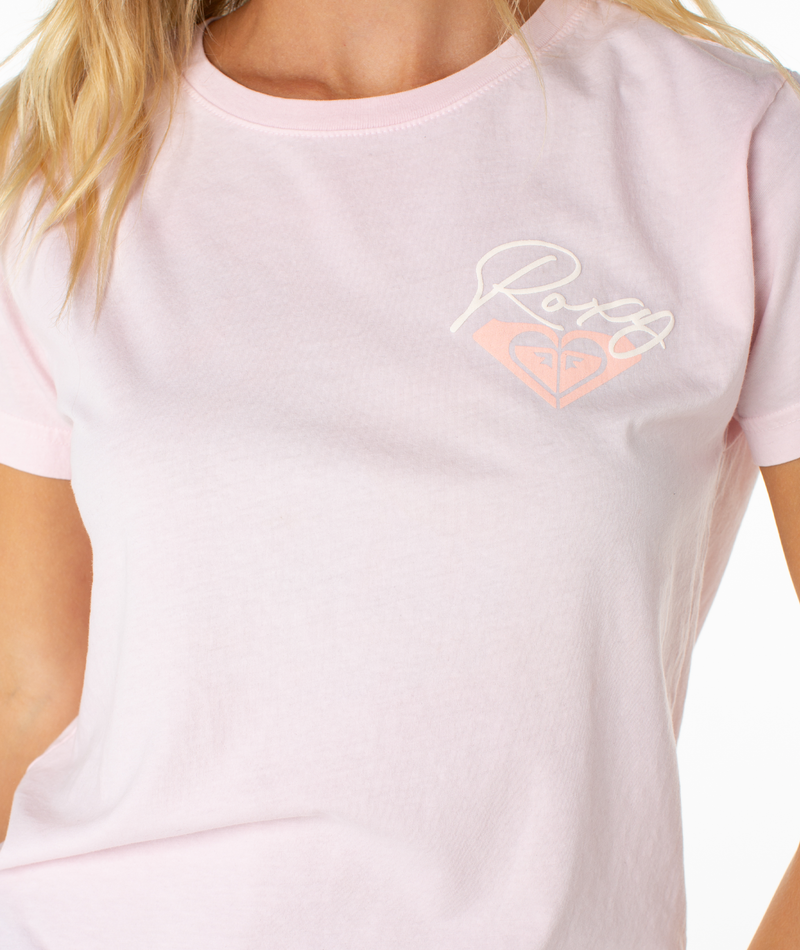 A person wearing the Roxy Monstera Circle Tee in light pink, featuring a white and peach logo with the brand name above a heart-shaped wave on the left chest. Only the upper torso is visible.