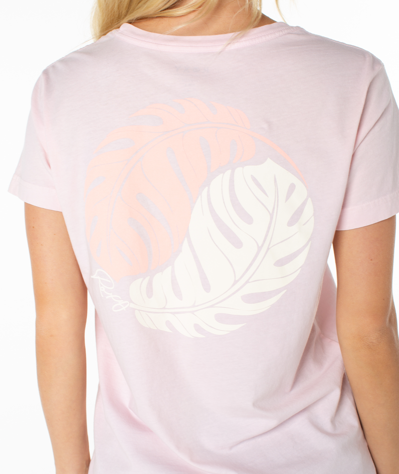 A person wears the Roxy Monstera Circle Tee, a light pink top featuring a large pastel graphic of two overlapping monstera leaves—one peach, one off-white—on the back. Only the back of the tee and part of their blonde hair are visible.