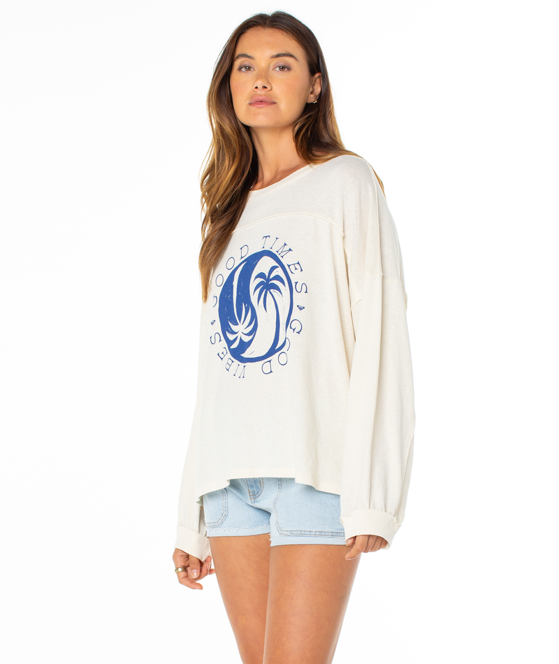 A woman with long brown hair wears the Roxy Changing Tides Top, a slouch pullover with balloon sleeves and a blue palm tree "GOOD TIMES GOOD VIBES" graphic, paired with light denim shorts, standing against a plain white background.