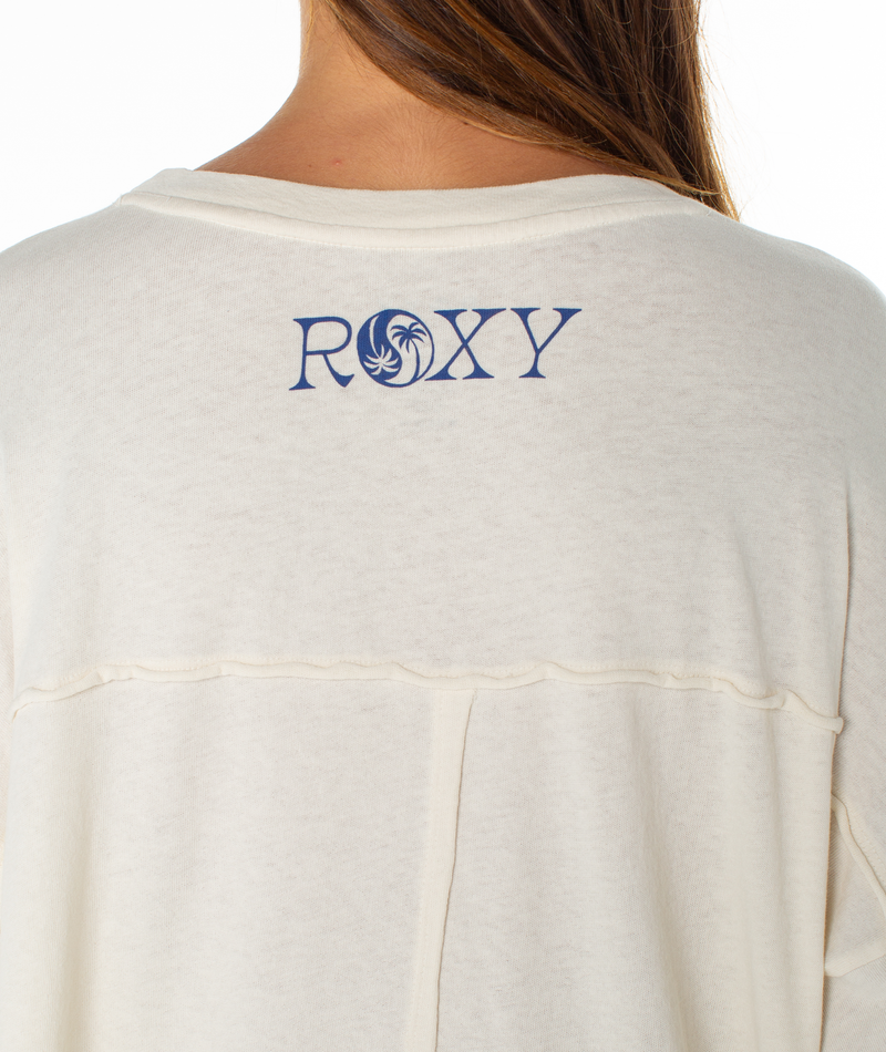 A woman with long brown hair wears an off-white Changing Tides Top by Roxy. The oversized shirt's back displays a blue ROXY logo featuring a palm tree inside the "O".
