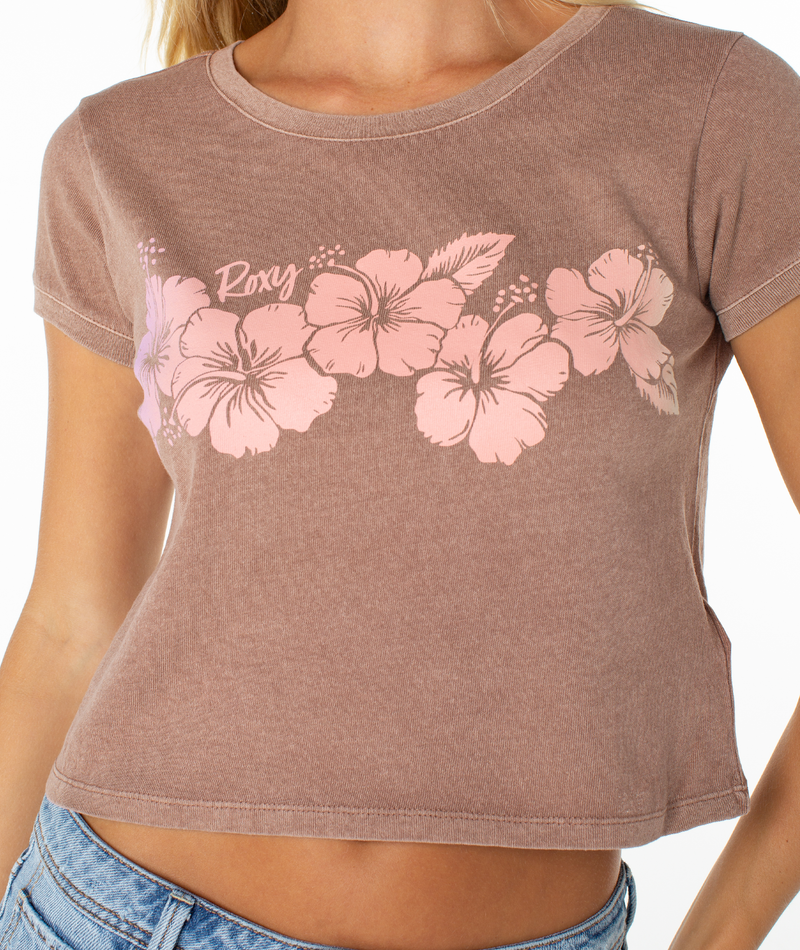 A woman wears the Roxy Hibiscus Row Tee—a light brown cropped t-shirt with pink hibiscus graphics and "Roxy" on the chest—paired with light blue jeans for an effortless summer look.