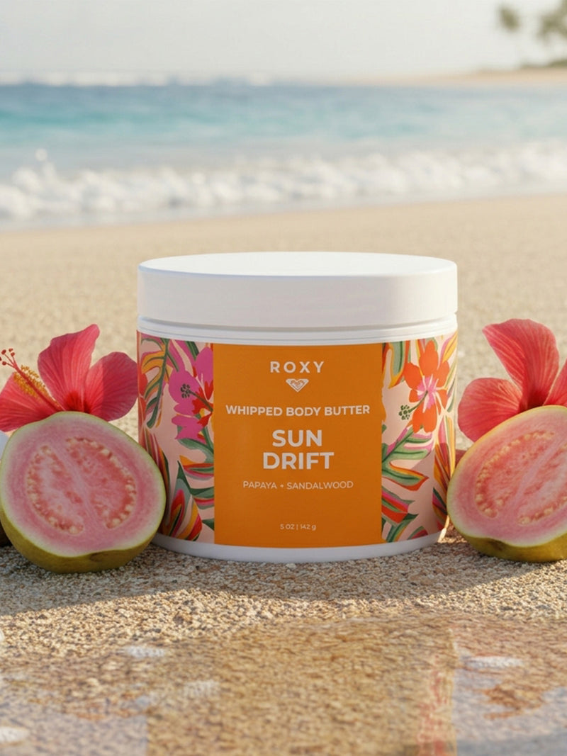 A jar of Roxy Sun Drift Body Butter, a hydrating lotion for dry skin, rests on sand beside halved guavas and red hibiscus flowers, with blurred ocean waves and palm trees in the background.