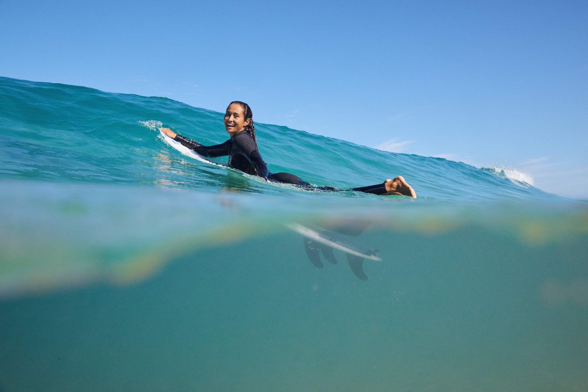 Surfer in a black ROXY wetsuit paddling through a turquoise ocean wave under a clear blue sky.