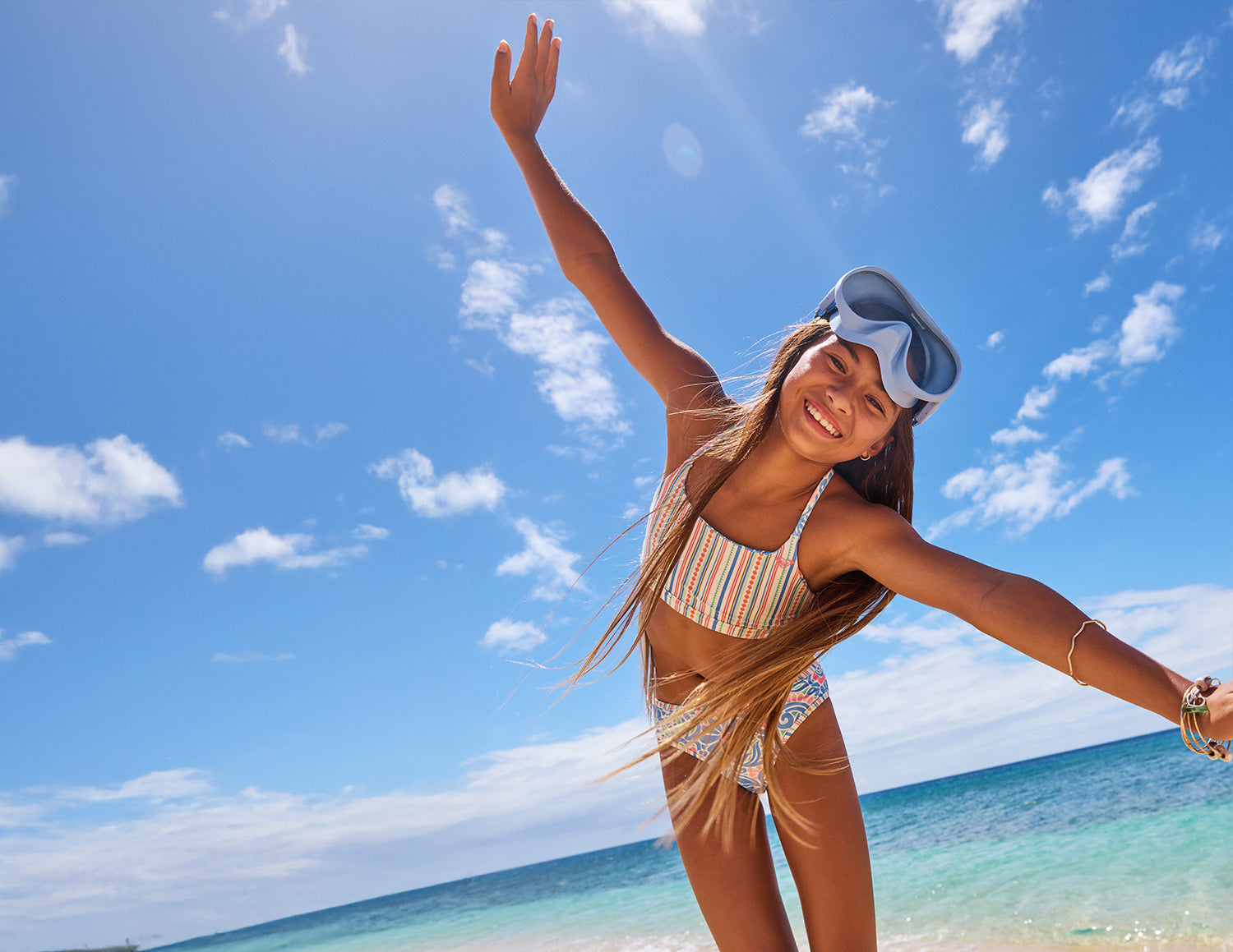Woman in a striped swimsuit and visor standing on a beach with clear blue sky and ocean.