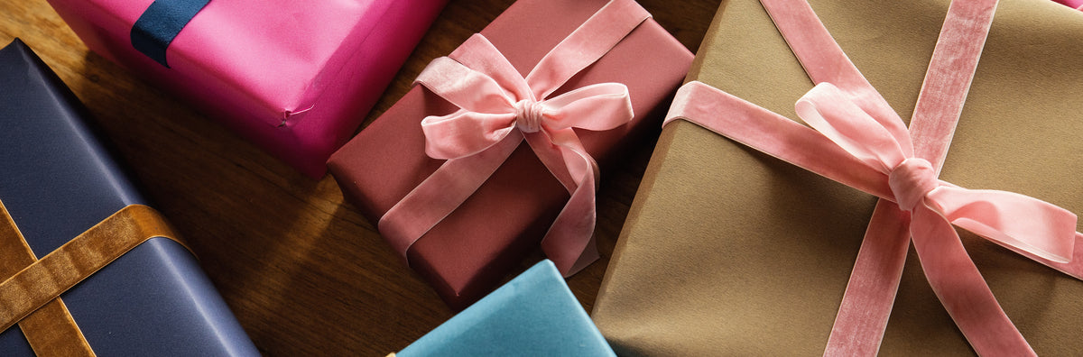 Several wrapped gift boxes arranged on a wooden surface, including pink, kraft brown, navy blue, and light blue packages with satin ribbon bows.