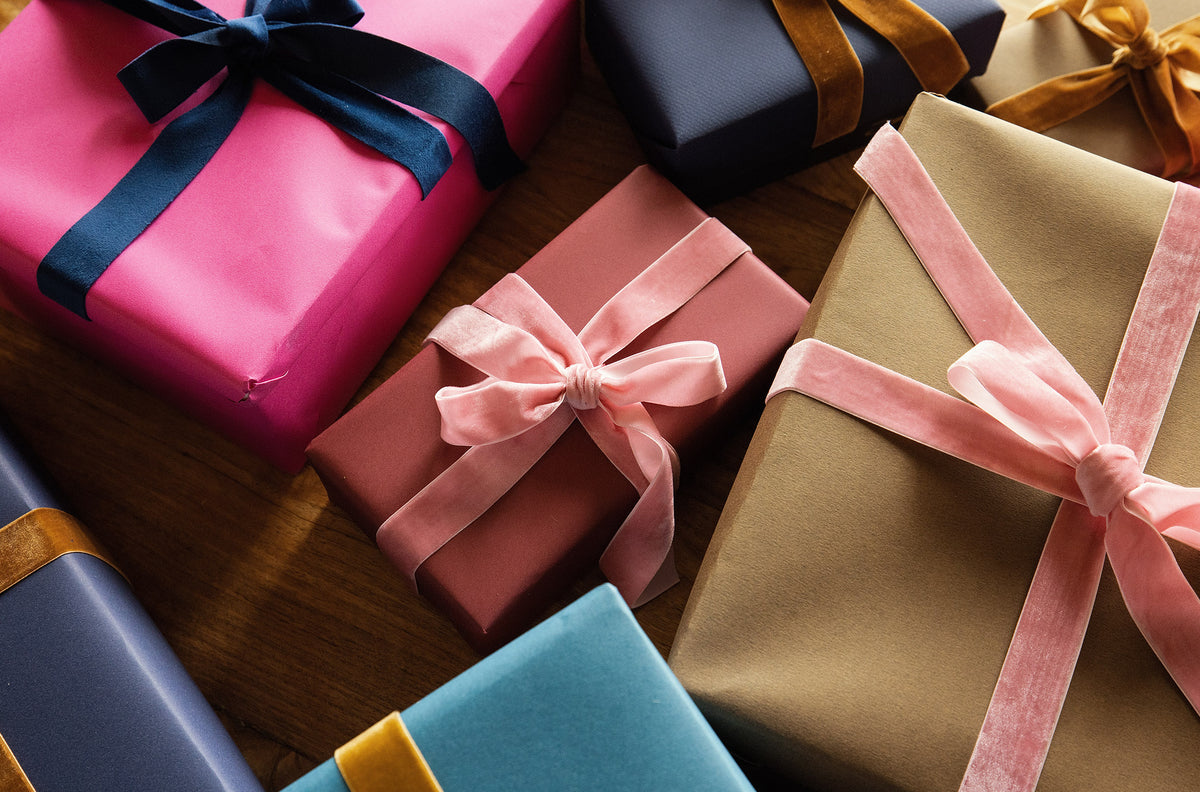 A collection of wrapped gift boxes in various colors, including pink, brown, blue, and gold, tied with ribbons and bows on a wooden surface.