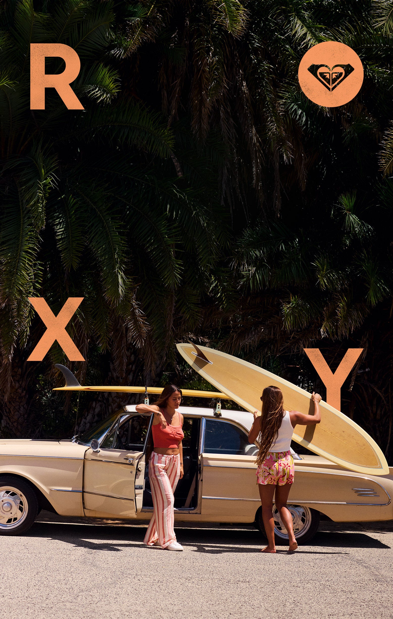 Two women with surfboards standing next to a vintage car, with 'Roxy' branding.