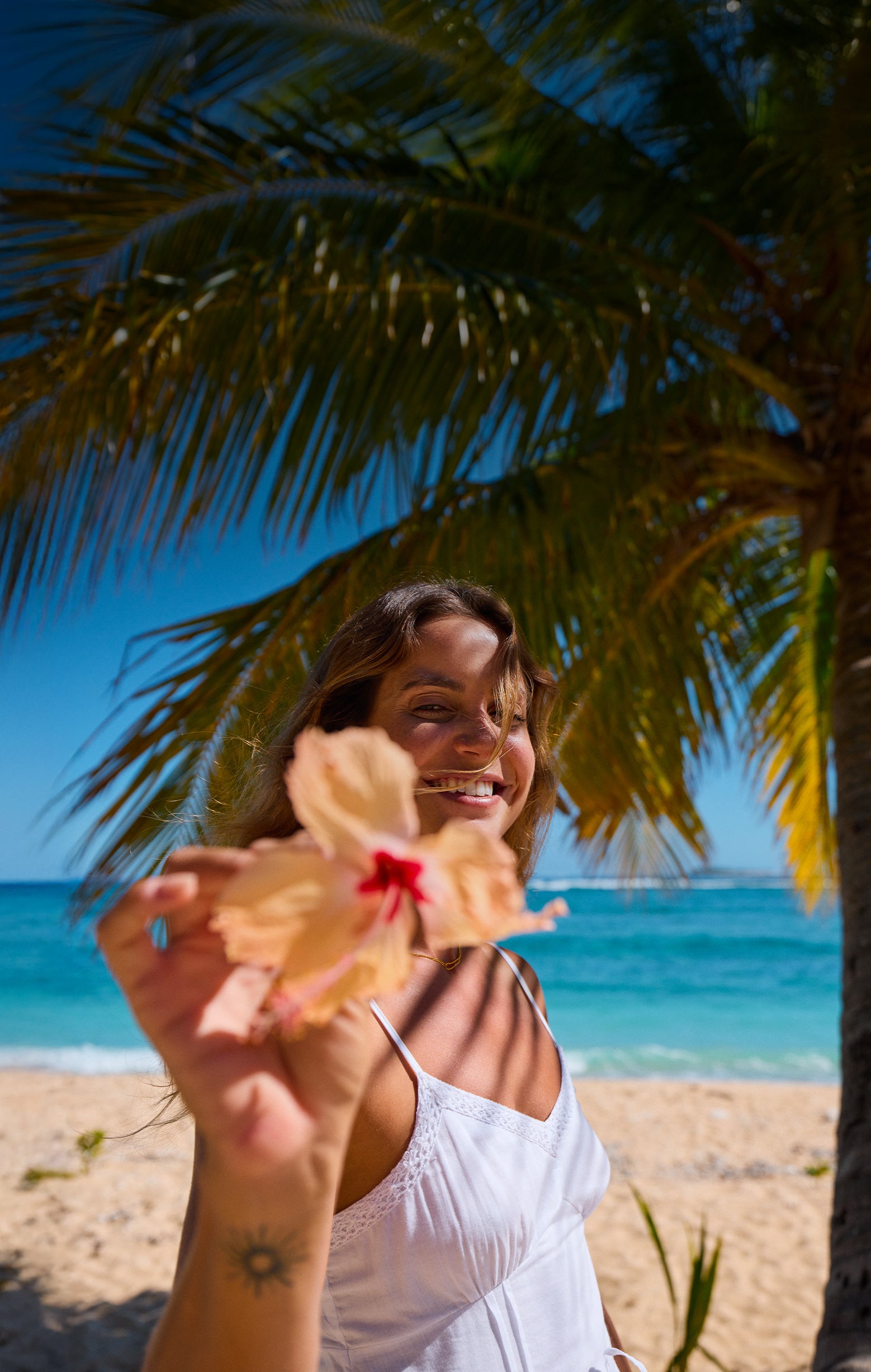 Woman holding a dried flower on a beach with palm trees and blue sky.