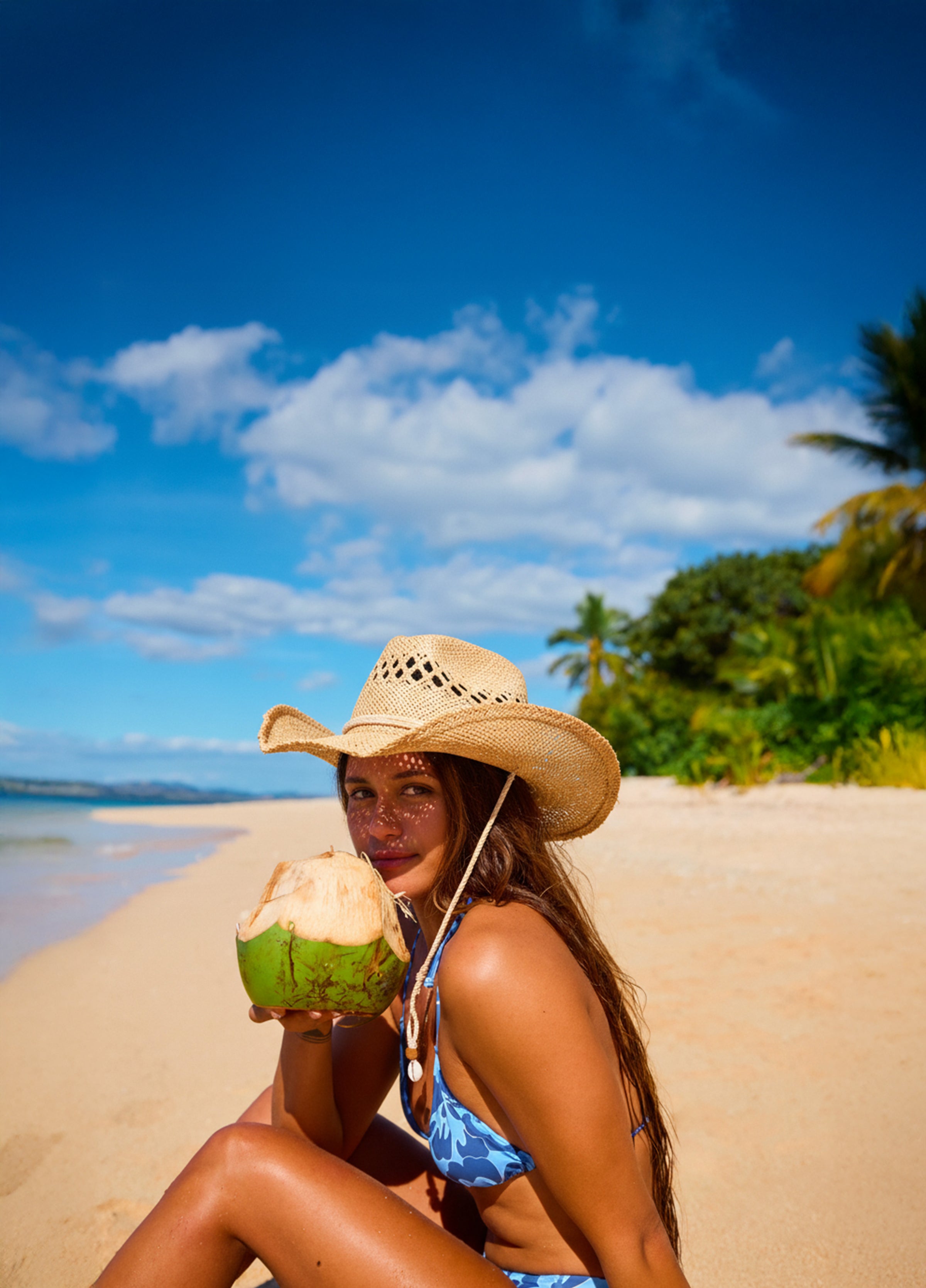 Woman in a bikini and hat holding a coconut on a beach with palm trees and blue sky.