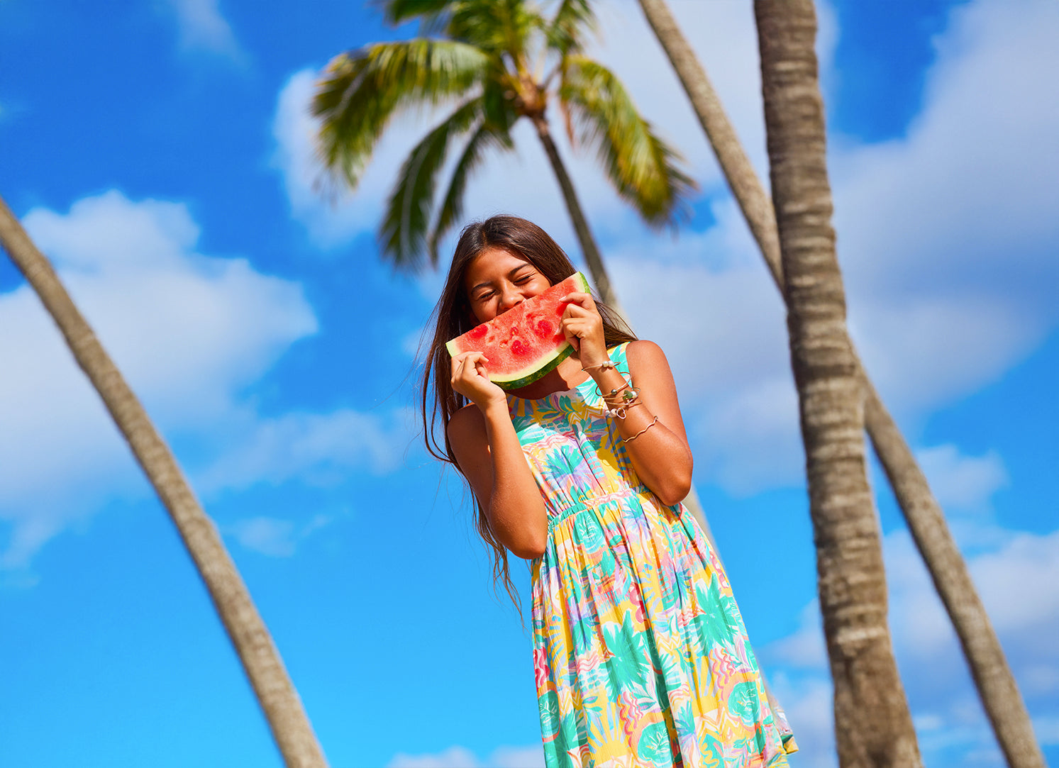 Woman in a colorful dress holding a watermelon slice with palm trees and blue sky in the background
