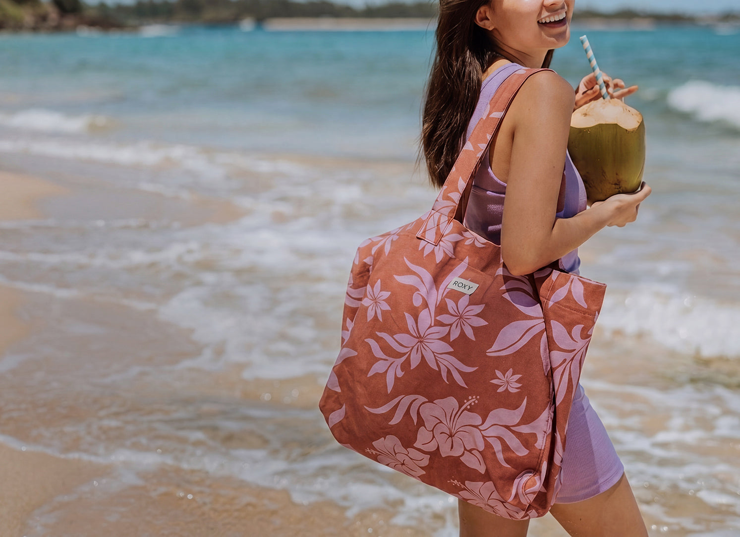 Woman on a beach holding a coconut and a floral tote bag