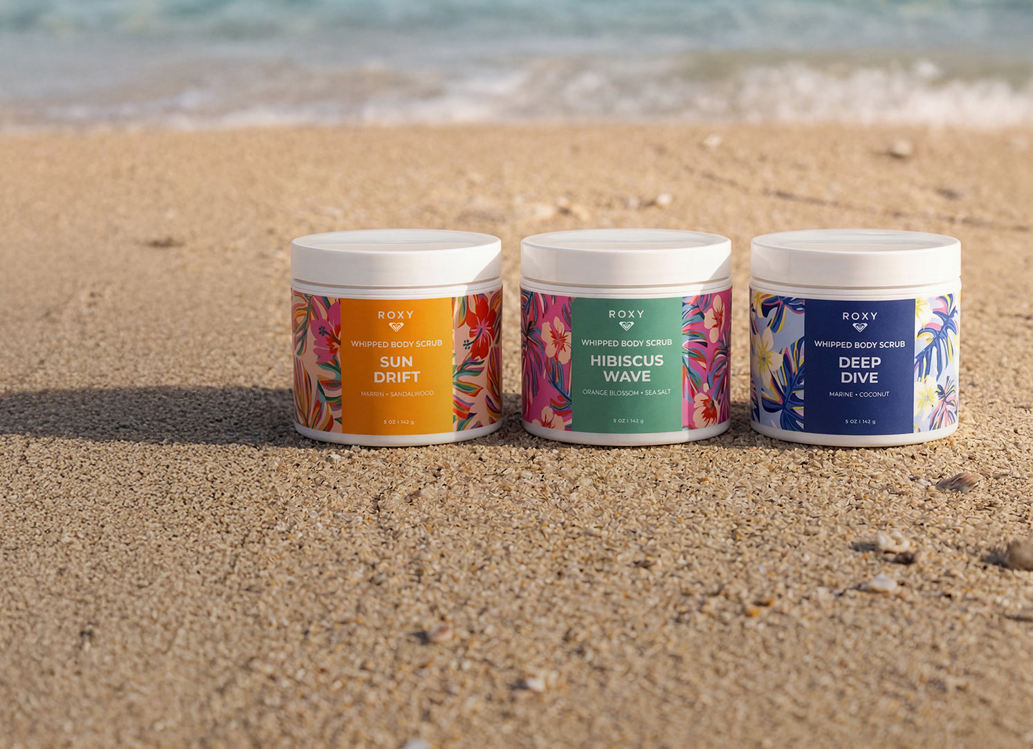 Three Roxy body scrubs on a sandy beach with ocean waves in the background