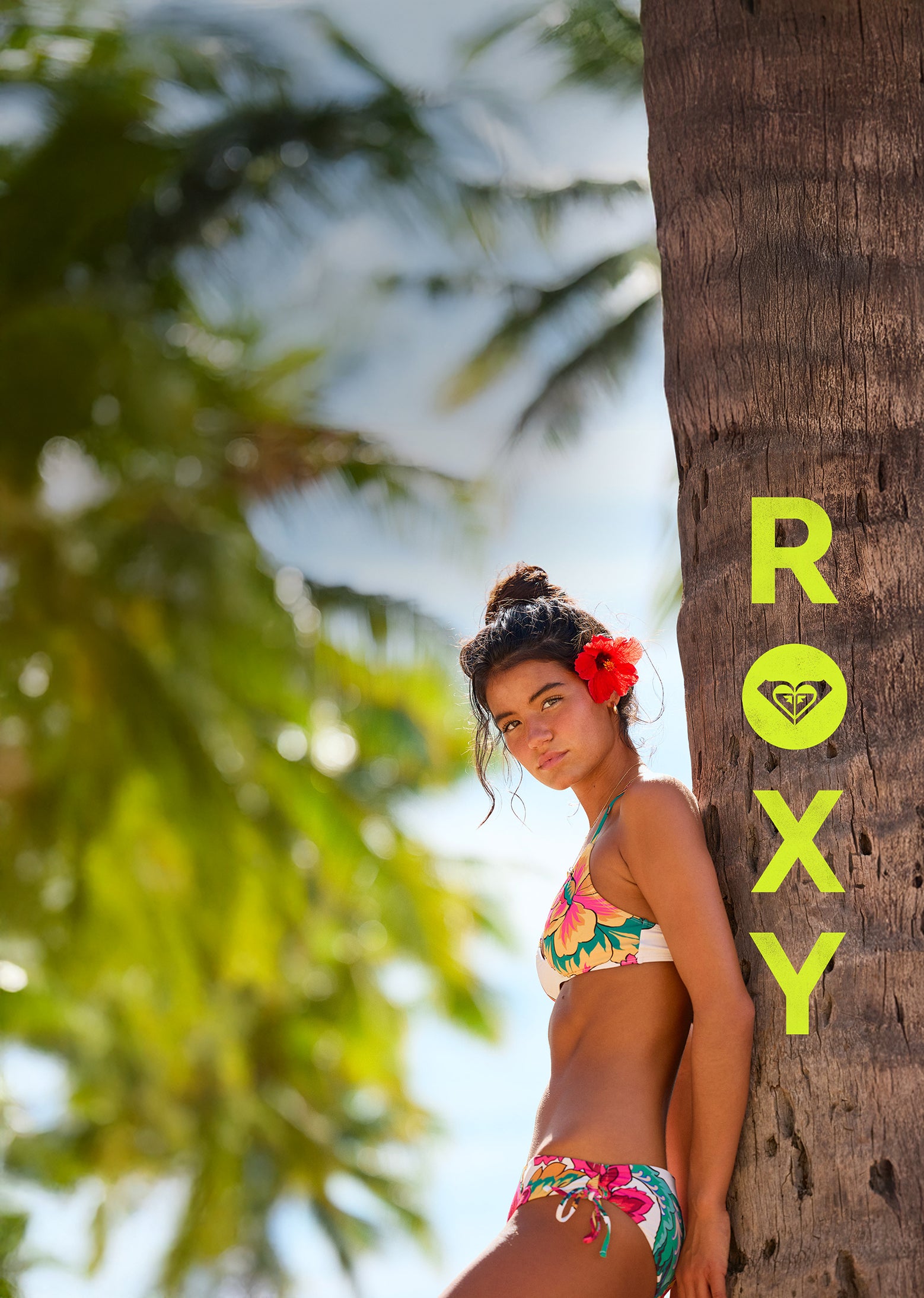 Woman in a colorful bikini leaning against a palm tree with 'Roxy' branding.