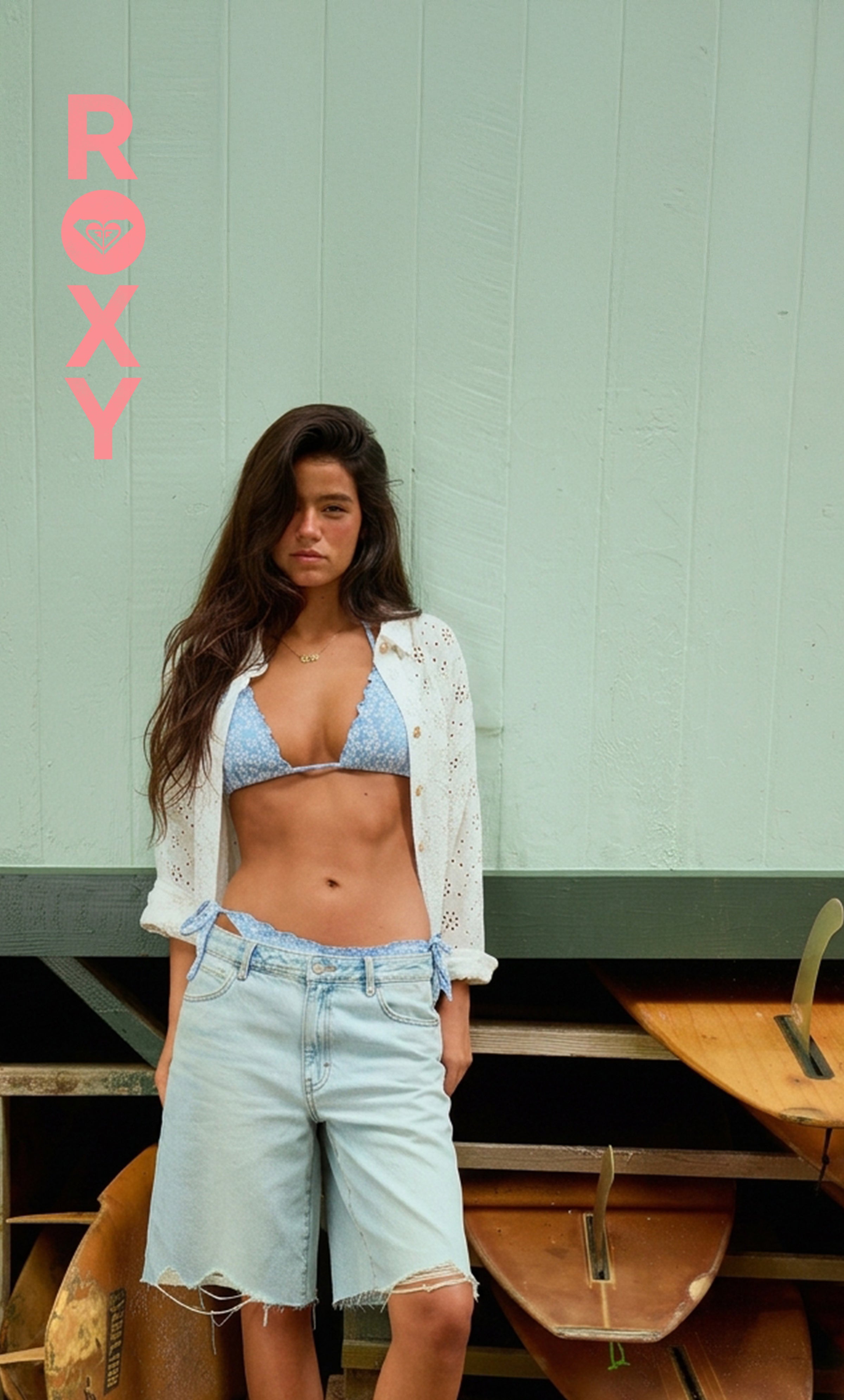 Woman in a bikini top and shorts standing in front of a green wall with 'Roxy' branding.