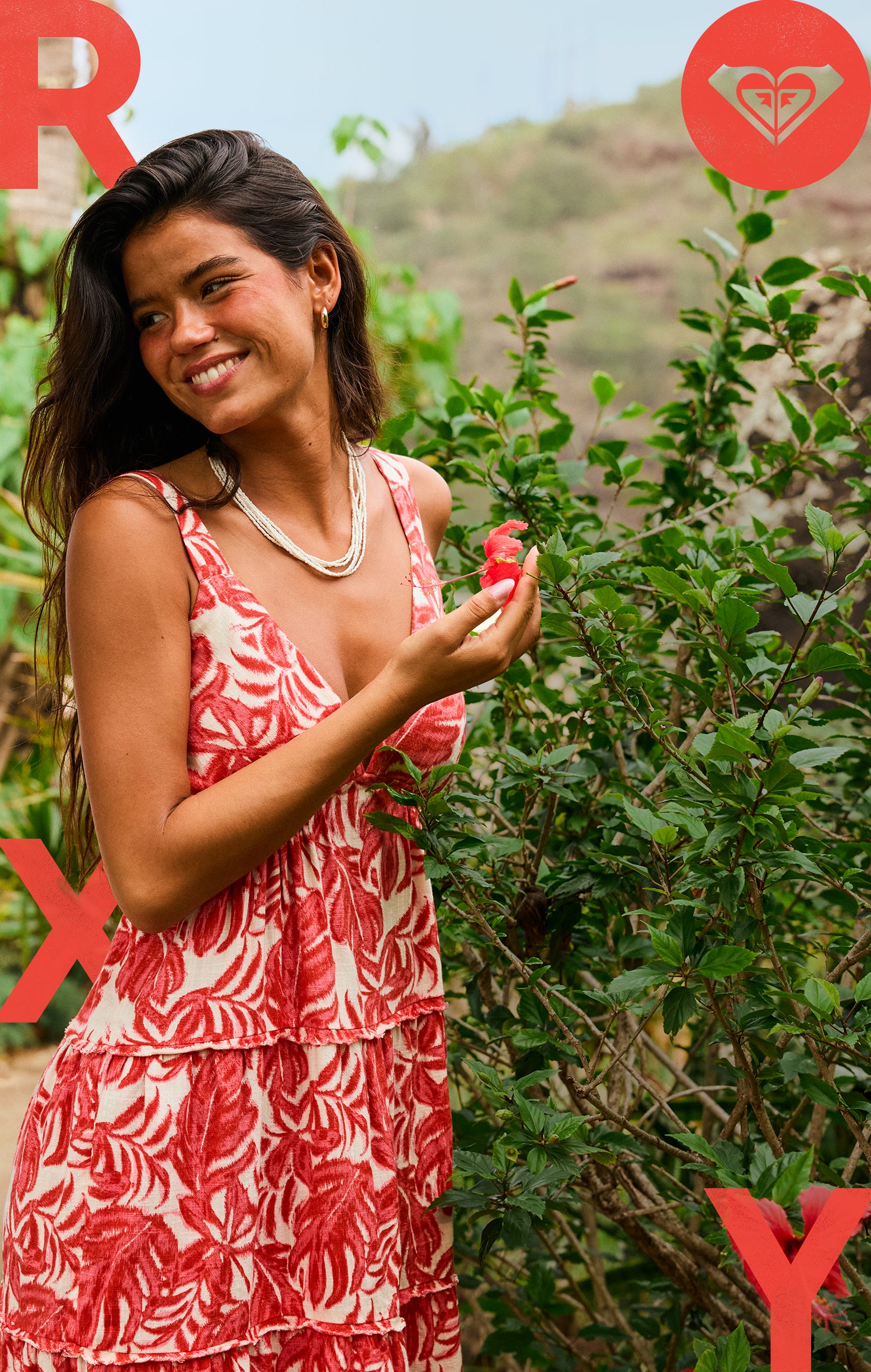 A woman in a red and white leaf-patterned dress stands smiling outdoors near green bushes, holding a red flower. The letters R and X and a heart-shaped logo appear in red on the image corners.