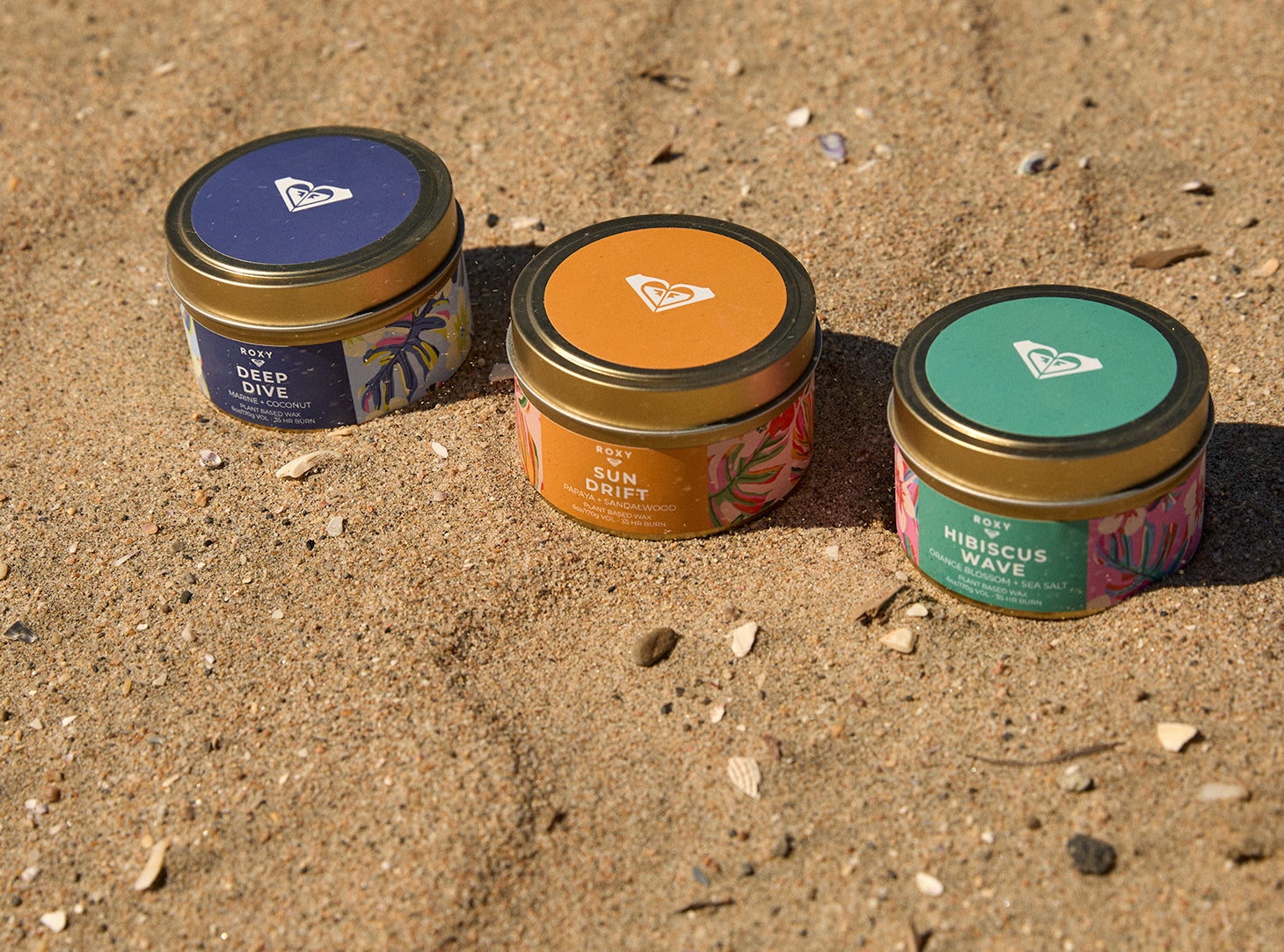 Three small round metal tins of fragrance or candle products arranged on sandy ground outdoors. The lids are dark blue, orange, and teal, each with a white heart logo and the text 'RIP' and 'DEEP DIVE', 'SUM DRIP', and 'HIBISCUS WAVE' respectively.