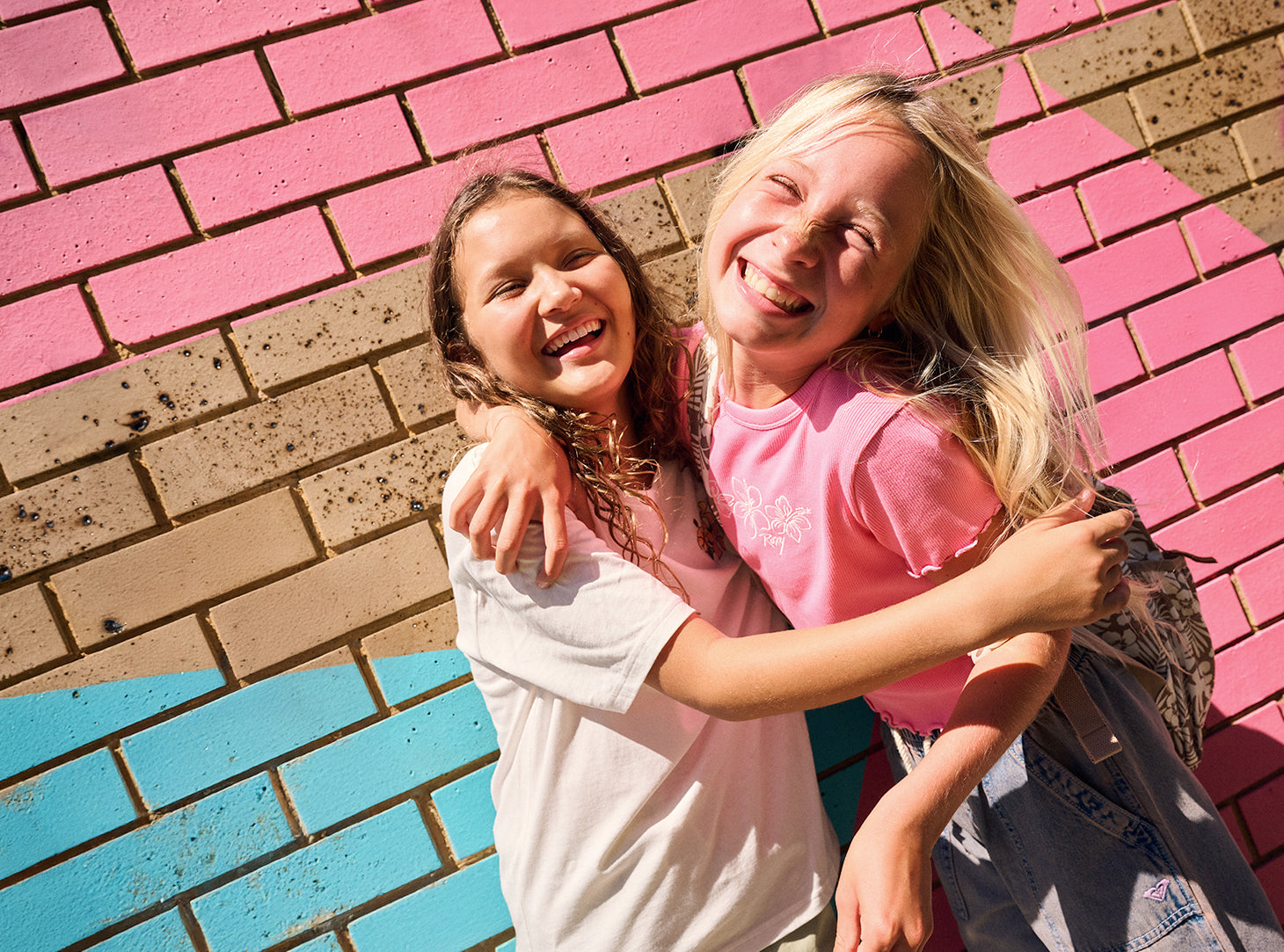 Two young girls hugging and smiling while wearing pink and white t-shirts, standing in front of a painted brick wall with pink, blue, and brown sections.