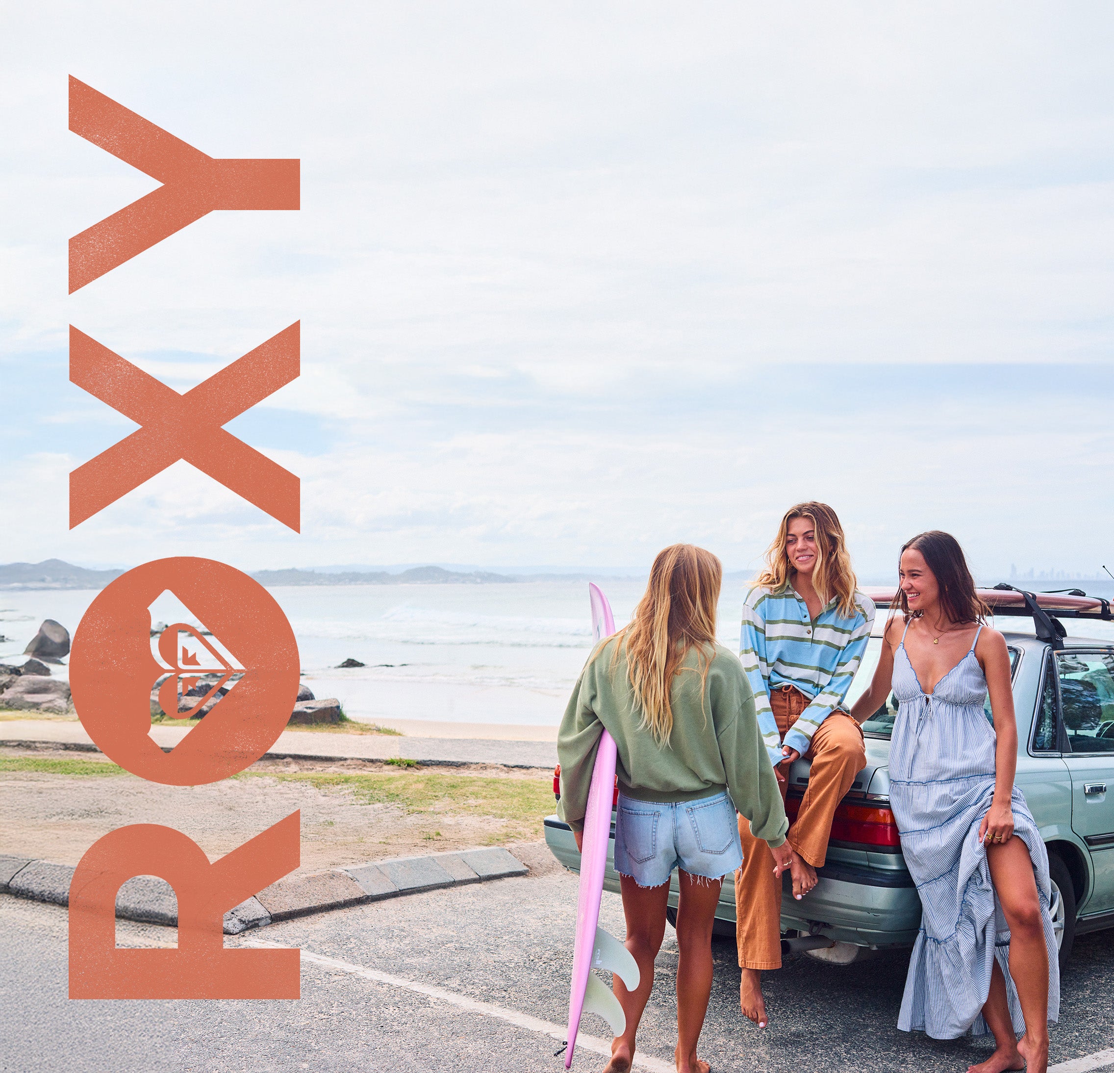 Three women standing beside a car on a beach road, with ocean and shoreline in the background. One woman holds a pink surfboard.