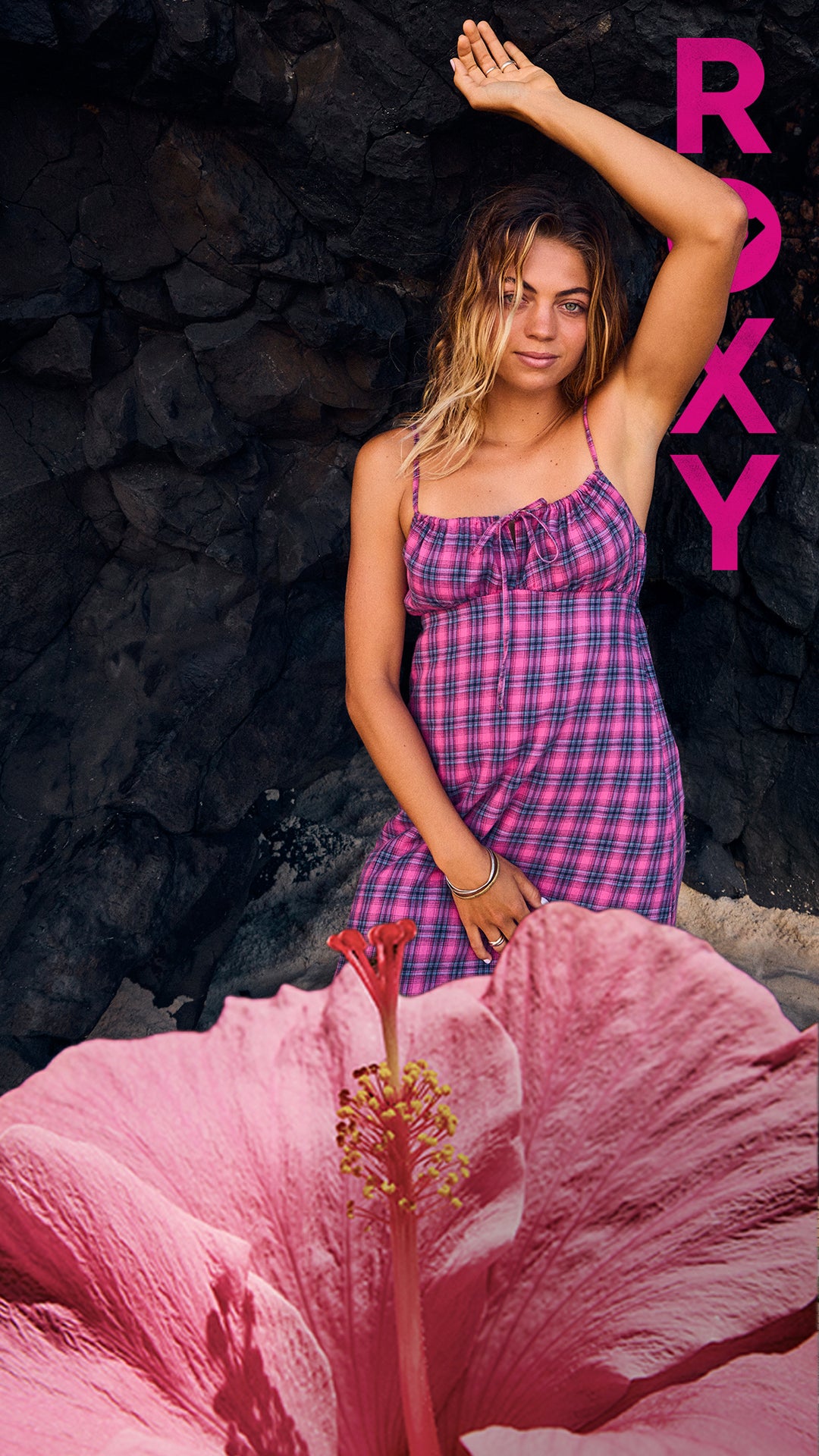 A woman wearing a pink and purple plaid spaghetti-strap dress stands against a dark rocky background, with a large pink hibiscus flower in the foreground and a vertical pink 'ROXY' logo on the right side.