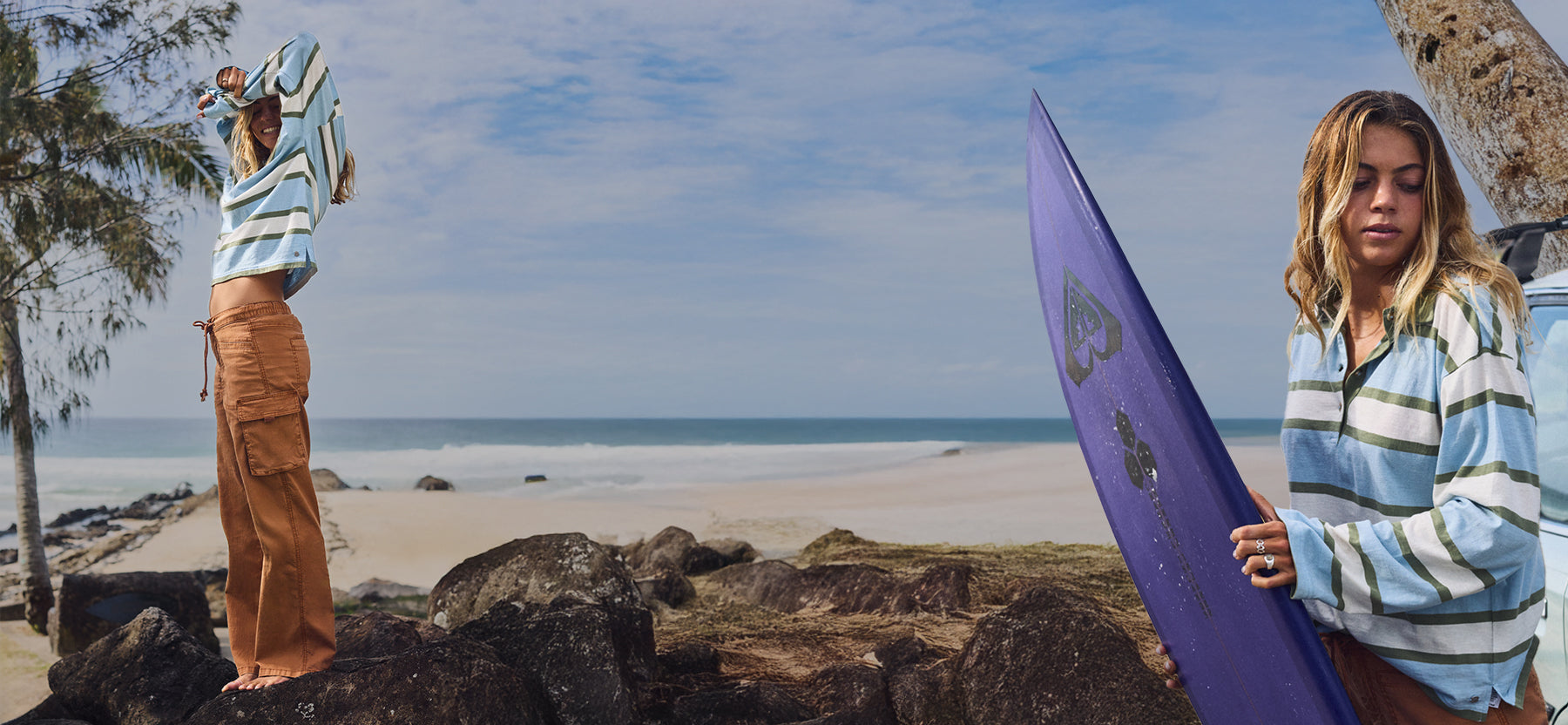 Two women on a beach wearing matching light blue and white striped long-sleeve shirts. One woman stands on rocks by the ocean wearing brown cargo pants, while the other holds a purple surfboard on the beach.