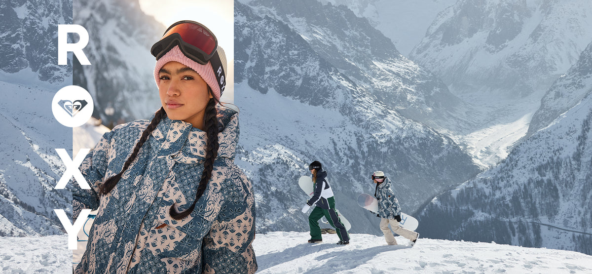 A snowy mountain scene with a woman in the foreground wearing a patterned ski jacket, pink beanie, and goggles, with two snowboarders further back. Large vertical Roxy branding appears on the left side.