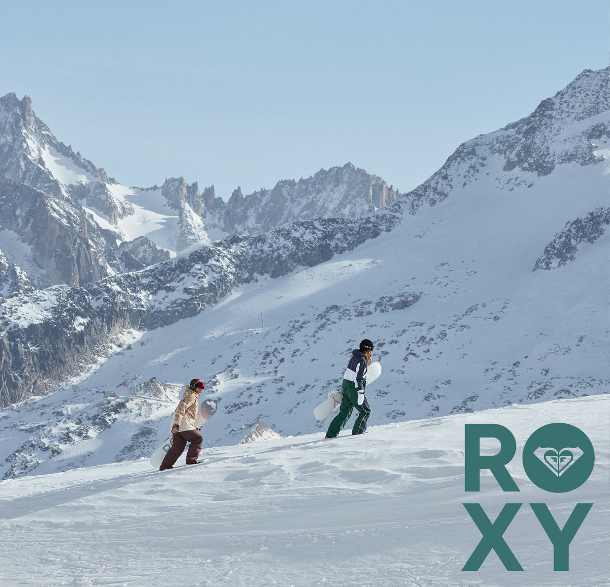 Two snowboarders on a snowy mountain slope with jagged snow-covered peaks and a pale blue sky. A large ROXY logo appears in the lower right corner.