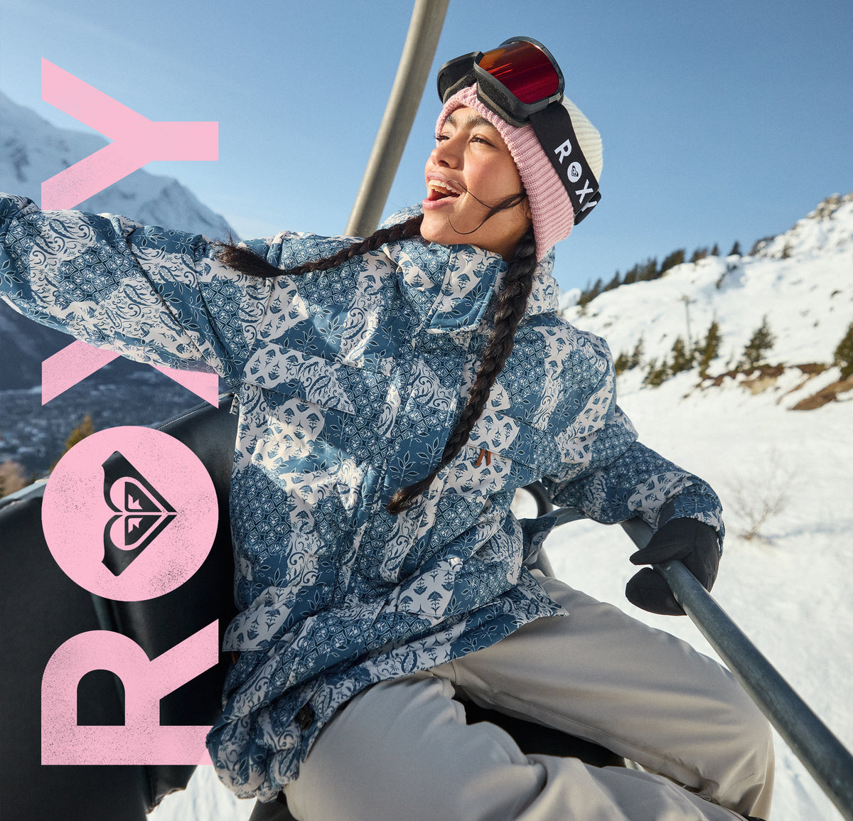 A woman wearing a blue and white patterned Roxy ski jacket, pink beanie, and ski goggles sits on a chairlift in a snowy mountain setting. Large pink Roxy branding is overlaid on the left side of the image.