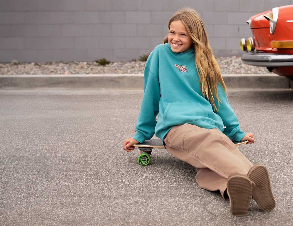 A young girl with long blonde hair sits on a skateboard on an asphalt surface, wearing a teal sweatshirt and beige pants. A gray brick wall and the rear of a red classic car are visible in the background.