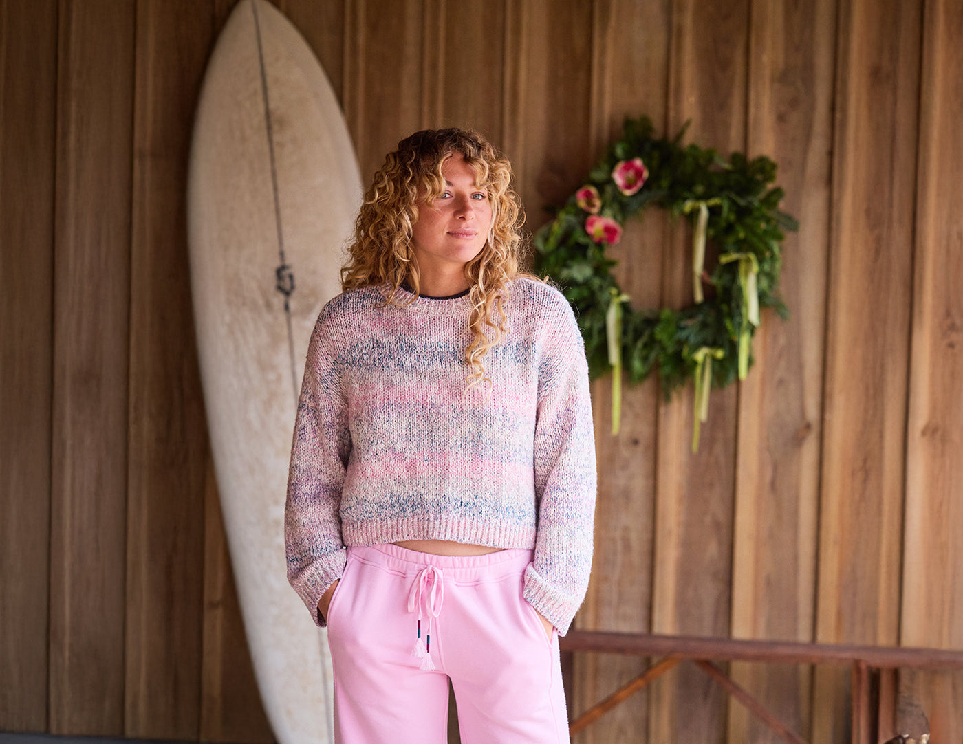 Woman in a colorful sweater and pink pants standing next to a surfboard against a wooden wall with a wreath.