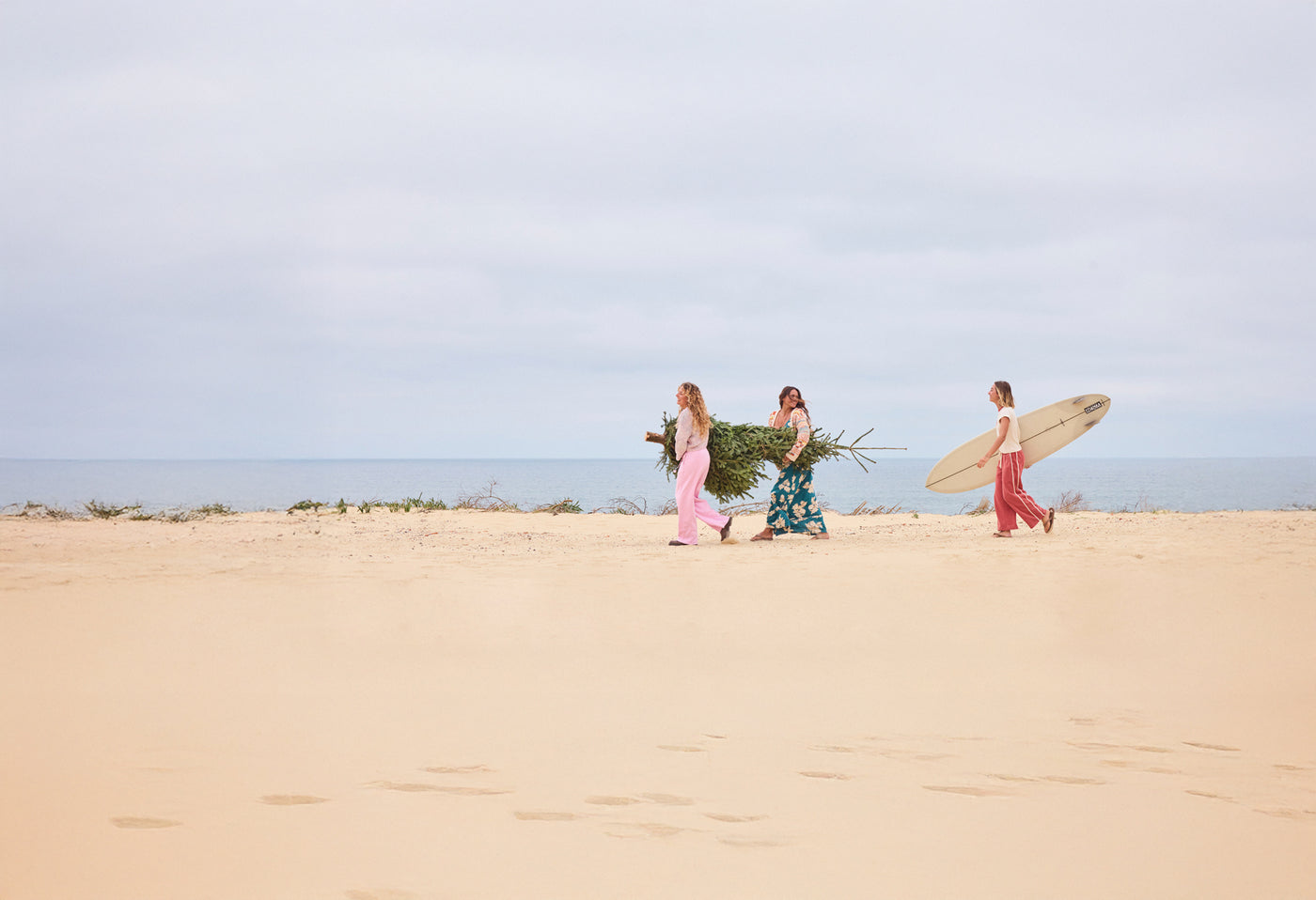 Three people walking on a sandy beach with surfboard and a tree
