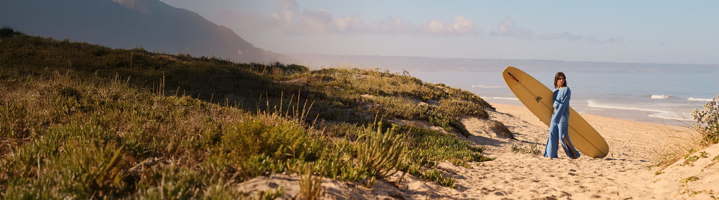 Person holding a surfboard on a beach with grassy dunes and ocean in the background
