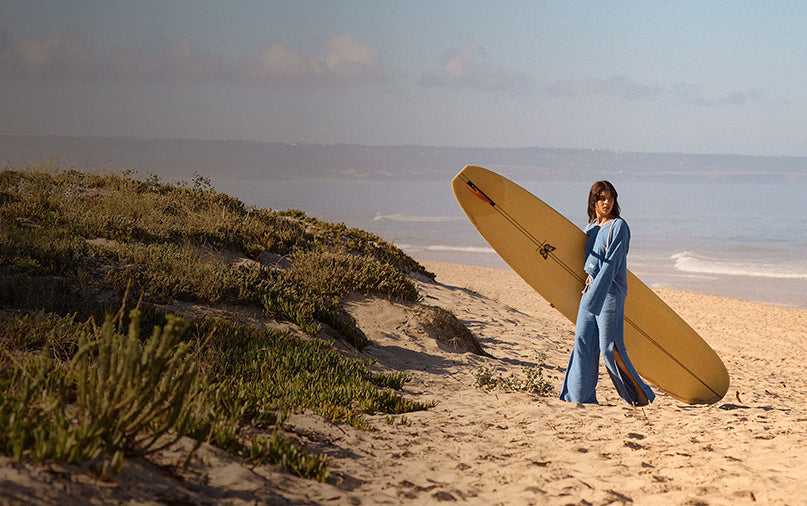 Person holding a surfboard on a sandy beach with grassy dunes and ocean in the background