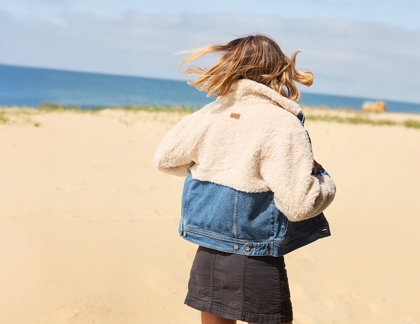 Person wearing a denim jacket over a beige fleece jacket on a beach