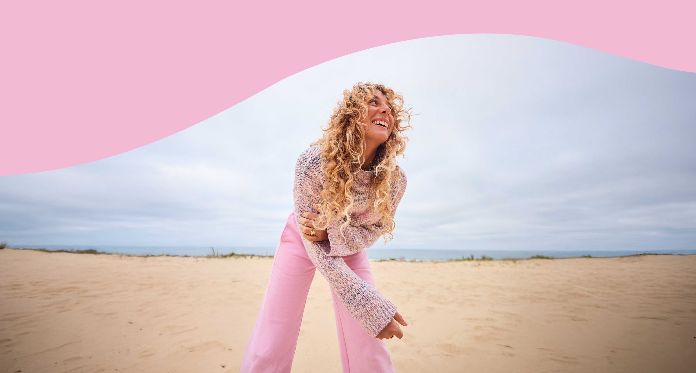 Person with long curly hair standing in a desert landscape with pink and blue abstract shapes.