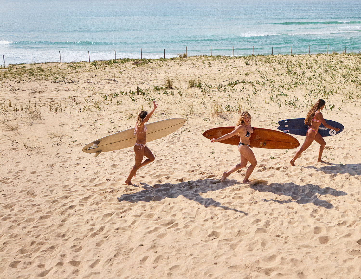 Three women walking on a sandy beach with surfboards.