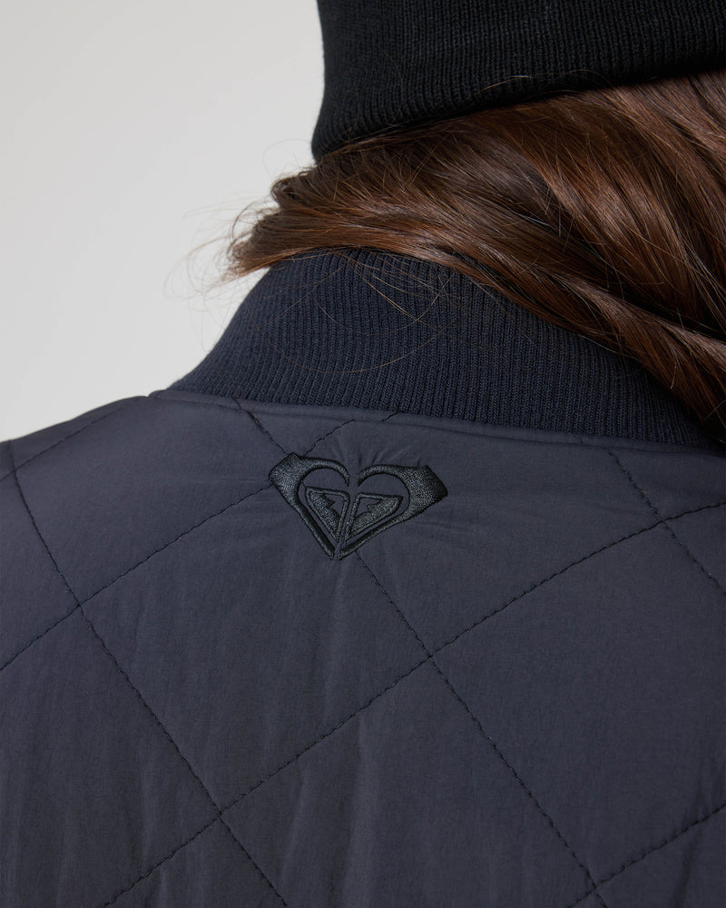 A close-up of a person’s back shows the Roxy Aurora Quilted Bomber featuring a stitched heart-shaped logo near the collar, with long brown hair peeking out from under a black beanie.