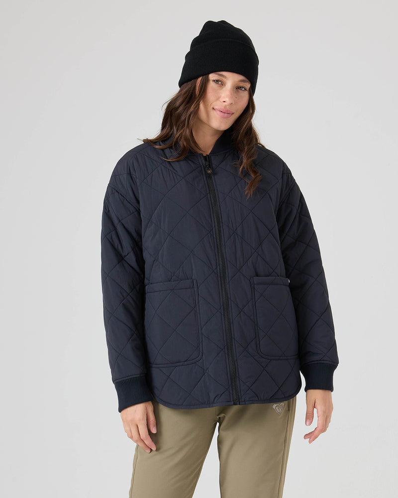 A person wearing a black beanie, khaki pants, and the Roxy Aurora Quilted Bomber jacket—featuring a crinkle nylon shell for added style and durability with front pockets in navy—stands against a plain light gray background.