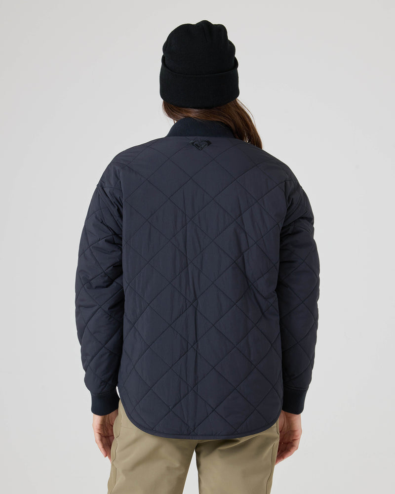 A person wearing a black beanie and the Roxy Aurora Quilted Bomber, a navy blue jacket with a diamond pattern crafted from a crinkle nylon shell, stands facing away from the camera. The jacket is paired with khaki pants against a plain white background.