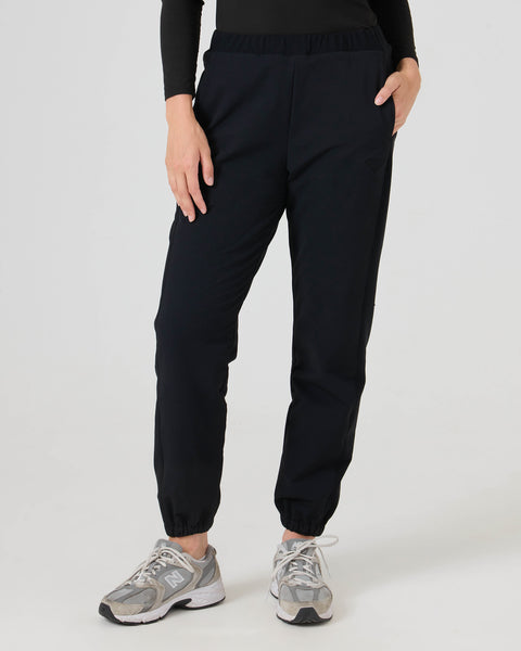 A person wearing Roxy’s Ember Textured Jogger in black, paired with a long-sleeve black top and gray New Balance sneakers, stands with one hand in their pocket against a plain light background.