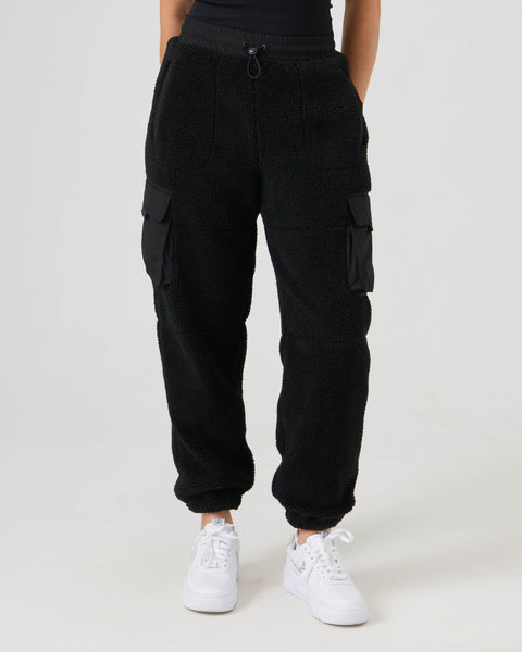 A person wearing the Roxy Rita Sherpa Jogger in black, featuring side pockets, paired with white sneakers, standing against a plain light background.