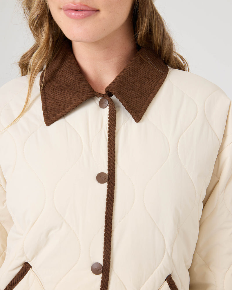 A woman wearing the Roxy Nyvea Quilted Barn Jacket, featuring a cream color with brown corduroy collar and trim. The image captures her from the shoulders to the mouth, emphasizing the jacket’s textured quilting and button details.