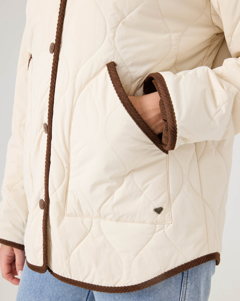 Close-up of a person wearing the Roxy Nyvea Quilted Barn Jacket—a cream jacket with brown trim and buttons—styled with blue jeans and their right hand tucked in the pocket.