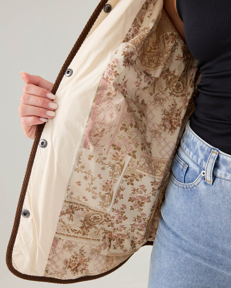 A person wearing jeans and a black top holds open the Roxy Nyvea Quilted Barn Jacket, revealing its cream lining with a pink and brown floral and geometric pattern.