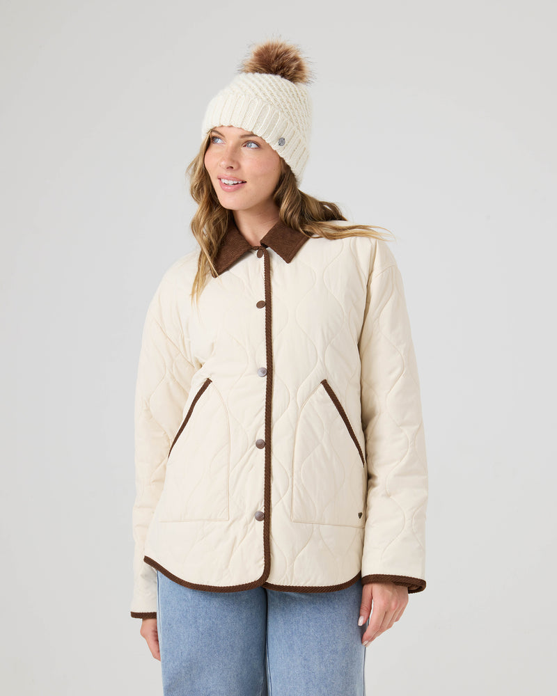 A woman wearing the Roxy Nyvea Quilted Barn Jacket in cream, featuring brown trim and buttons, paired with light blue jeans and a cream knit beanie with a brown pom-pom, stands against a plain light background.